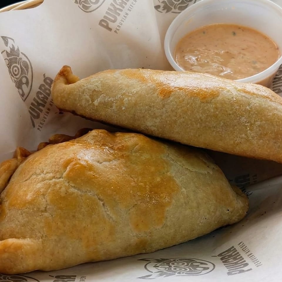 Two golden-brown Peruvian empanadas served with creamy dipping sauce on Pukará Peruvian Kitchen branded paper.