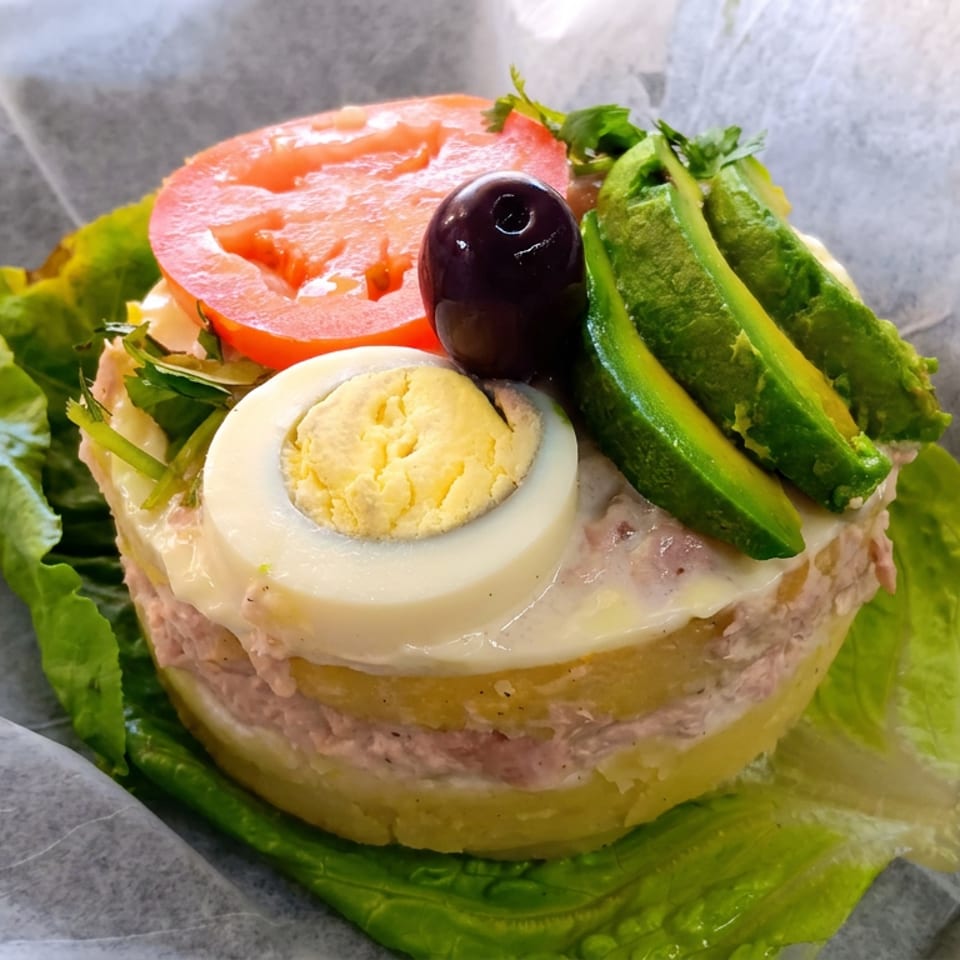 Layered savory dish on lettuce, topped with sliced tomato, cucumber, hard-boiled egg, black olive, and herbs.